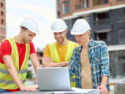 Three construction workers in hard hats and safety vests review plans on a laptop at a construction site, under the watchful eye of their Safety Supervisor, ensuring compliance with WSH standards.
